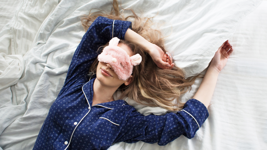Woman lying on a white bed sheets wearing blue pyjamas and a pink fluffy face mask